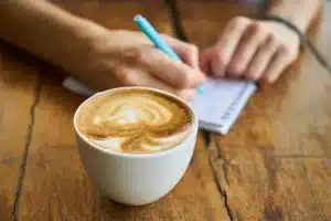 Hände auf einem Holztisch neben einem Notizbuch und einer Kaffeetasse vor einem Fenster